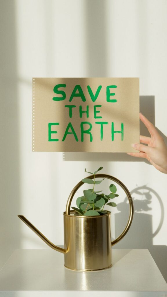 A hand holds a sign reading 'SAVE THE EARTH' above a brass watering can with a eucalyptus plant on a white background.