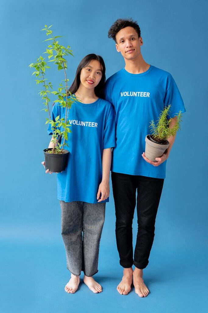 Two volunteers in blue shirts holding potted plants against a blue background.